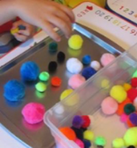 Student sorting pom poms of various sizes and colors into a bucket and tray.